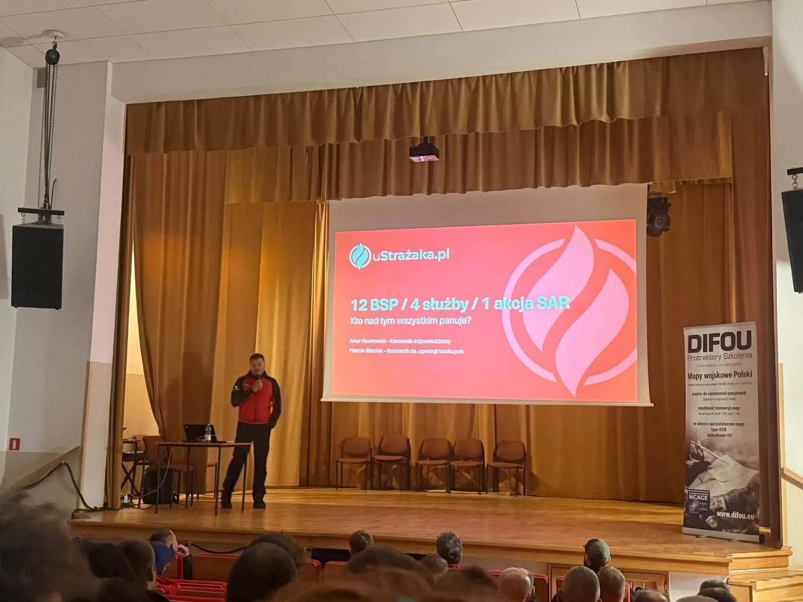Sala szkoleniowa pełna ratowników, na scenie prelegent uStrażaka.pl, na ekranie slajd „12 BSP / 4 służby / 1 akcja SAR. Kto nad tym wszystkim panuje?”